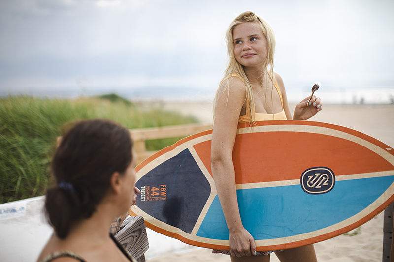 En tjej på en strand som äter glass med en surfbräda under armen.