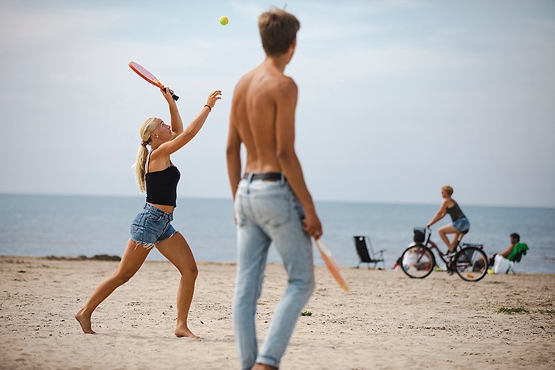 Ungdomar spelar tennis på stranden. I bakgrunden cyklar en person. 